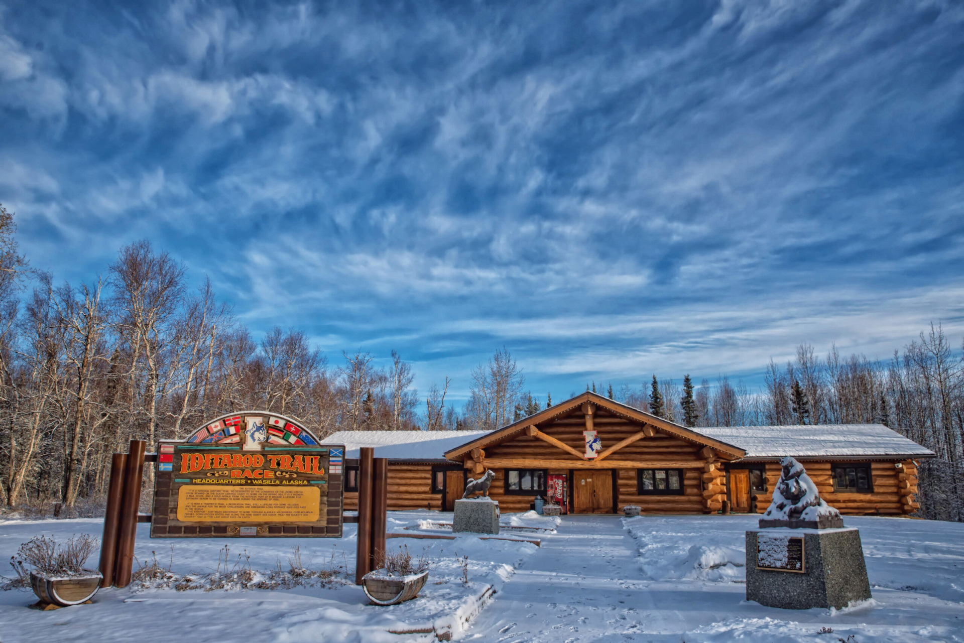 A log cabin building with a sign in front of it, surrounded by snow and trees under a blue sky.