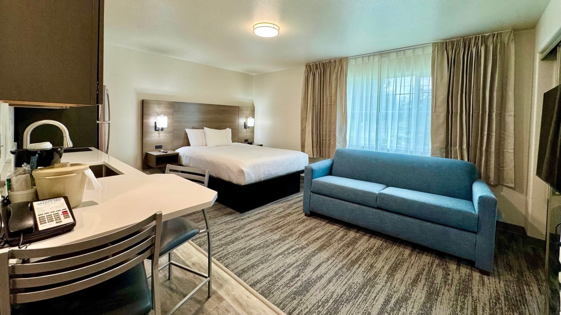 A hotel room with a kitchenette, sofa, bed, and TV.